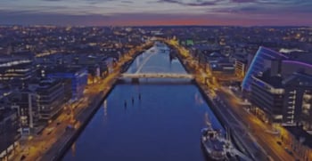 Aerial view of a cityscape at dusk with a river running through the center, illuminated bridges, and buildings lit up on both sides.