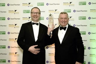 Two men in tuxedos smiling, holding an award at a formal event backdrop.