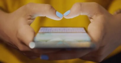 Hands with blue nail polish type on a smartphone, displaying a blurred keyboard. The person wears a yellow top, conveying focus and engagement.