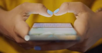 Hands with blue nail polish type on a smartphone, displaying a blurred keyboard. The person wears a yellow top, conveying focus and engagement.
