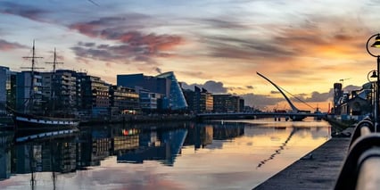 A tranquil river scene at sunset, reflecting colorful clouds and modern buildings along the waterfront, with boats docked nearby.