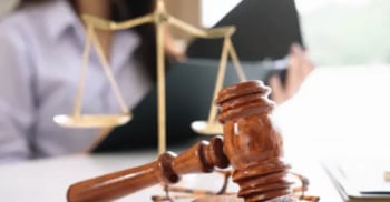 Judge’s gavel and scales of justice on desk, person reading legal documents in background.