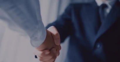 Two people are shaking hands, both wearing suits. The image conveys professionalism and agreement, with a softly blurred background.