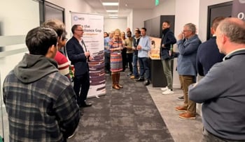 A group of people gathered in a hallway, with one person speaking at the front and others listening. There are banners and a poster in the background.