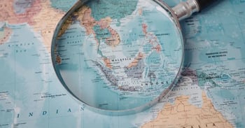 A magnifying glass is placed over a map focusing on Southeast Asia, highlighting countries like Malaysia, Indonesia, and surrounding regions.