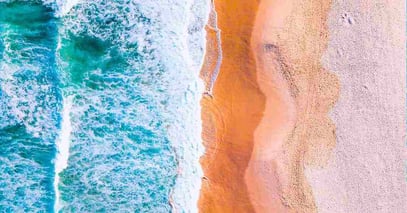 Aerial view of a vibrant beach scene with turquoise waves crashing onto an orange sand shoreline, creating a striking contrast. The mood is serene and colorful.