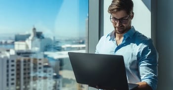A man with glasses smiles while using a laptop near a large window overlooking a cityscape, exuding a professional and focused atmosphere.