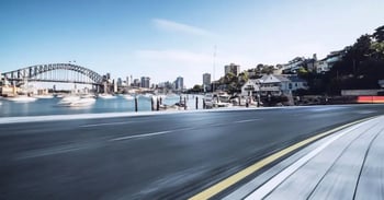 A serene urban scene with a winding road leading to a harbor. Boats move on calm water near an arch bridge under a clear blue sky.