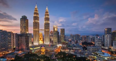 The Petronas Twin Towers and surrounding city buildings in Kuala Lumpur are illuminated at dusk under a partly cloudy sky, symbolizing a city where regulatory compliance is rising rapidly in Malaysia.