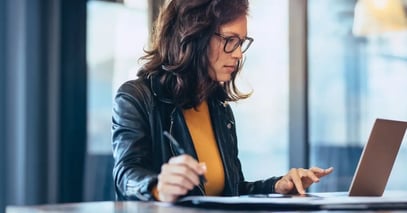 Woman wearing glasses working on laptop and taking notes at a desk in office.