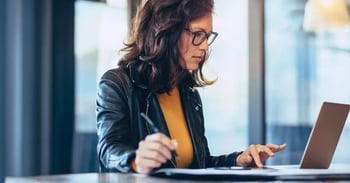 Woman wearing glasses working on laptop and taking notes at a desk in office.