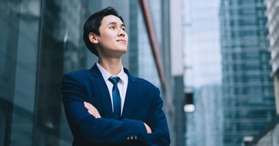Young professional in a blue suit stands confidently with arms crossed, looking up amidst tall modern skyscrapers, conveying ambition and optimism.