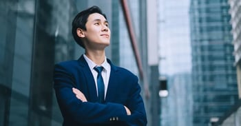 Young professional in a blue suit stands confidently with arms crossed, looking up amidst tall modern skyscrapers, conveying ambition and optimism.