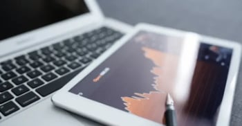 Tablet displaying financial graph beside laptop and pen on desk.
