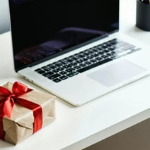 A wrapped gift with a red ribbon sits on a white desk next to an open laptop.