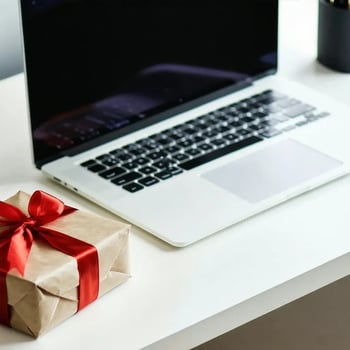 A wrapped gift with a red ribbon sits on a white desk next to an open laptop.