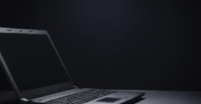 A closed laptop sits on a dark surface against a black background, with soft lighting highlighting the device from above.
